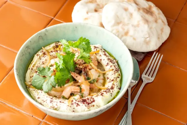 A bowl of hummus with prawns and coriander beside two pita breads on orange tiles