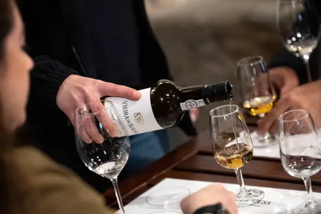 A hand pouring white wine from a bottle into a tasting glass on a wooden table