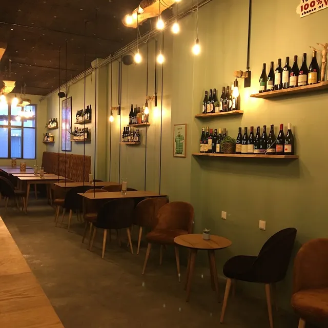 A long room with a green wall, wine bottle shelves and hanging bulbs above the tables