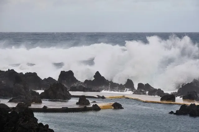 Water pools among black lava rocks facing a huge breaking wave