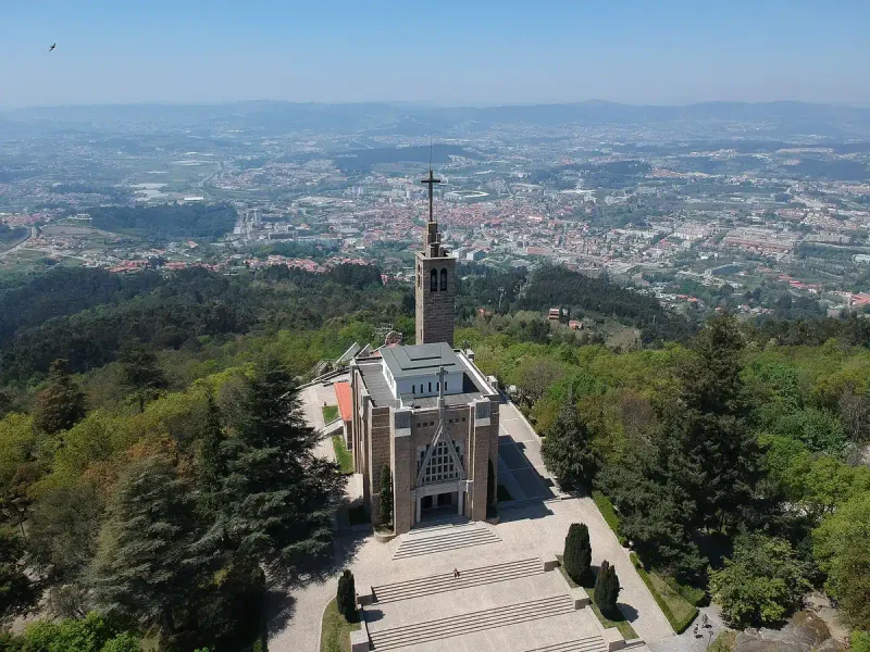 A stone church with a cross tower on a wooded summit, with a city spread out in the valley behind
