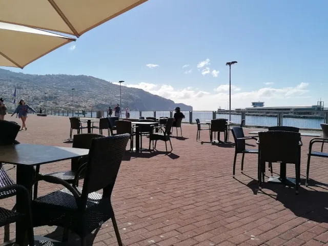 An empty promenade terrace with a parasol, dark chairs and a view of the sea and hills