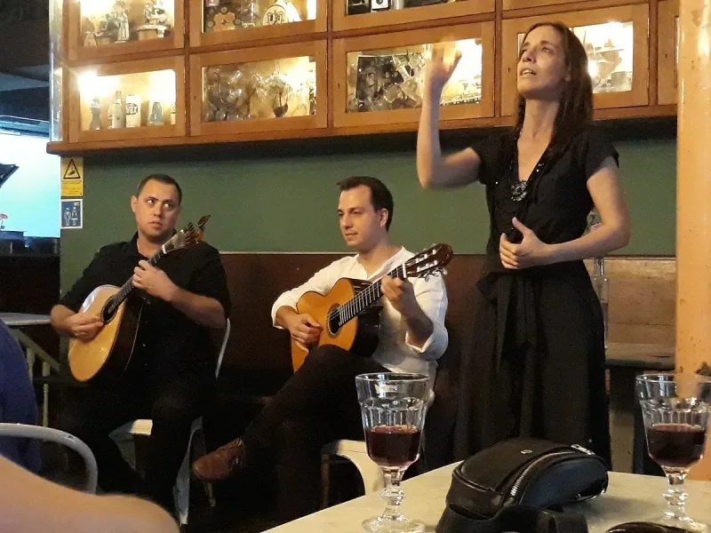 A fado singer in black mid-song, a Portuguese guitar player and a viola player seated beside her, and two glasses of red wine on the table in front