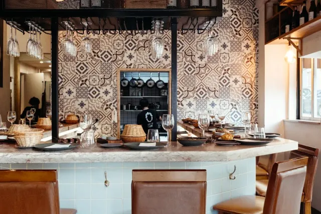 A marble counter facing a patterned tiled wall, hanging glasses and leather chairs