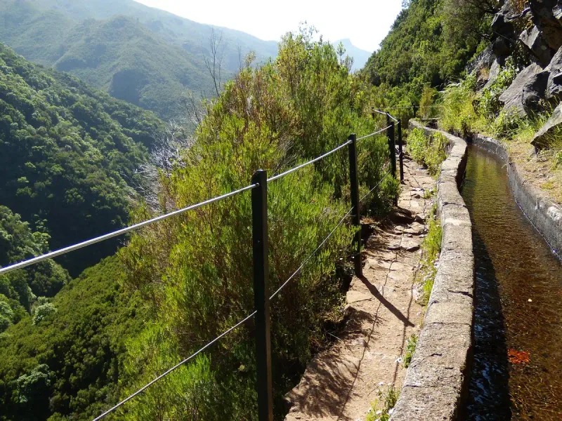 A narrow path with a water channel and a guide cable along a steep green mountainside