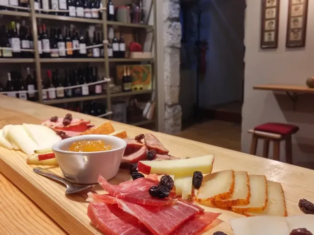 A board of cured meats, cheeses and jam on a wooden counter facing wine bottle shelves