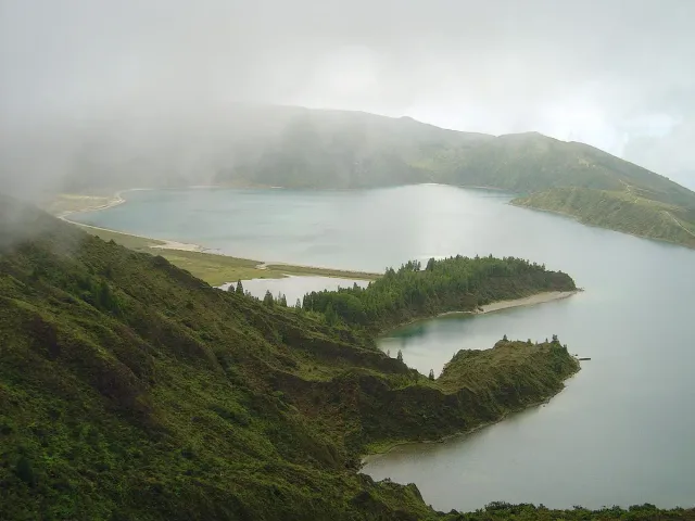 A crater lake with a pale sand strip between green ridges and haze