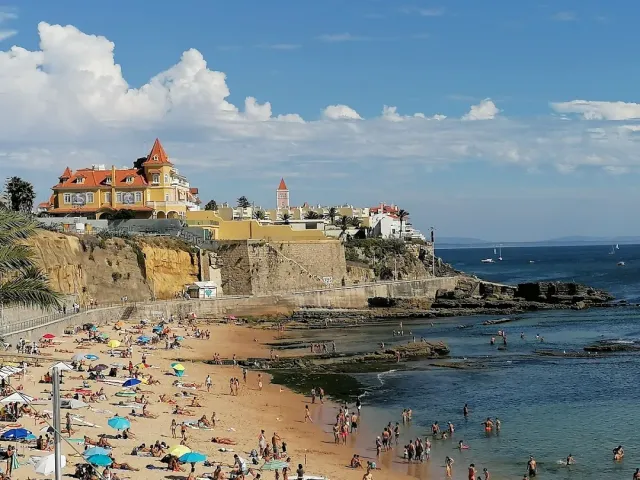 A small sandy bay full of bathers, with a yellow mansion and red turret above it