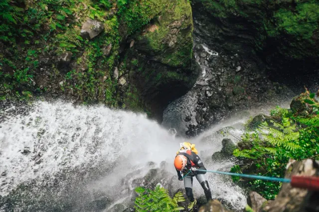 A person in an orange helmet rappelling beside a waterfall among green rocks