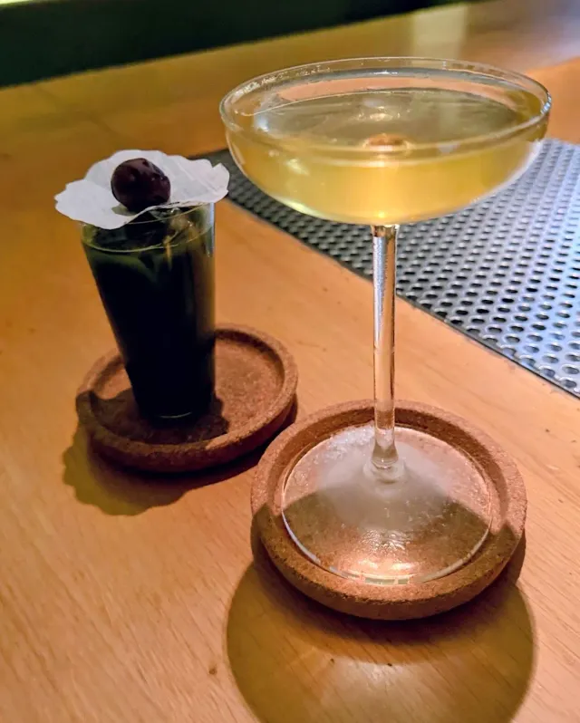Two cocktails on cork coasters, one dark and one yellow in a coupe
