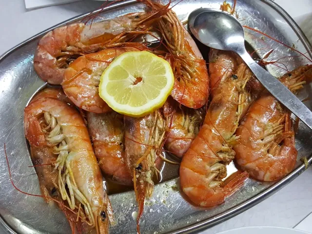 A metal tray with large grilled shrimp, a lemon slice and a spoon