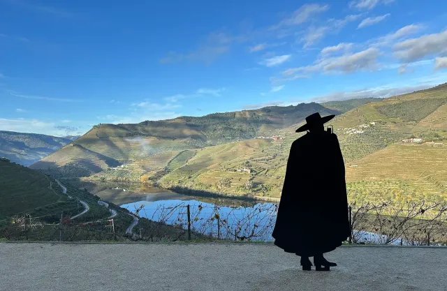 The silhouette of a caped figure in a wide hat facing a river bend between vineyard slopes