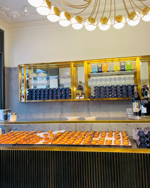 A counter with dozens of custard tarts on trays, brass-framed shelves and a gilded chandelier