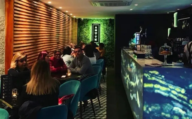 A dim space with a green plant wall, a lit sign, turquoise chairs and a glowing counter