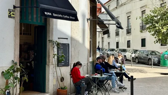 A street corner with a black awning, people sitting at small tables and parked cars
