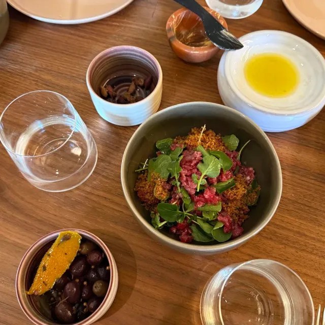 A bowl of tartare with green leaves on a wooden table, beside olives and olive oil