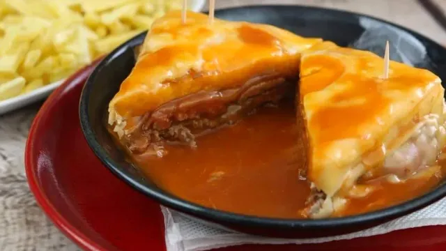 A layered sandwich cut open in a black bowl with orange sauce and fried potatoes