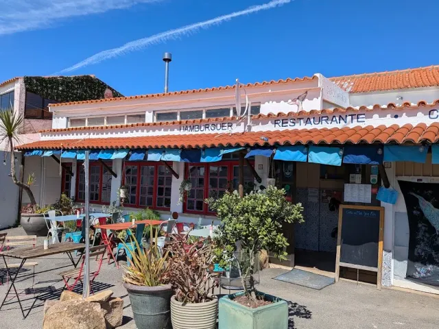 A white building with roof tiles, blue awnings, colourful chairs and potted plants in the sun
