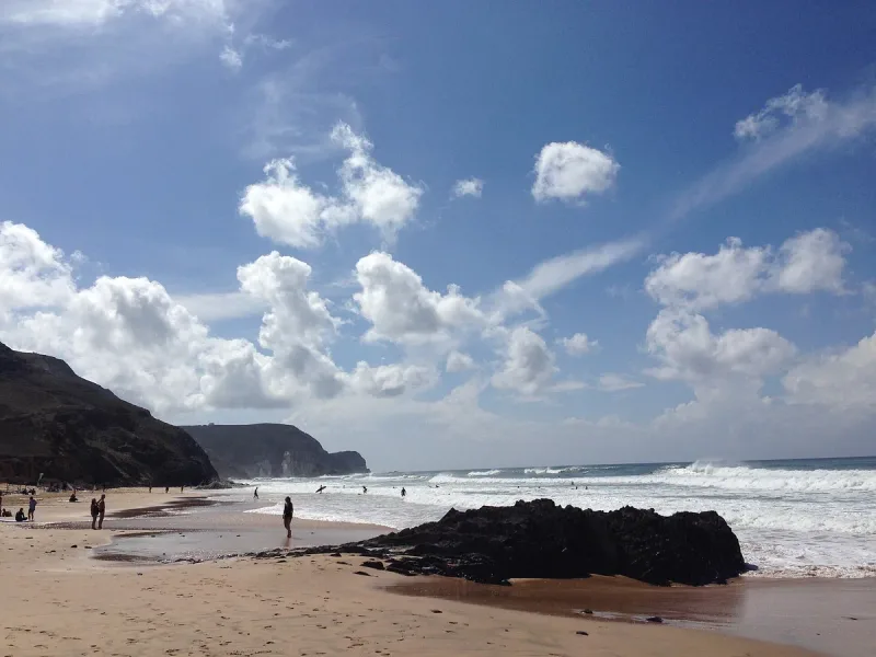 Wild sandy beach with a black rock outcrop and breaking surf beneath a blue sky and white clouds