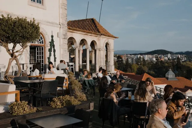 A terrace with diners beside a stone building with arches and a view of the city roofs