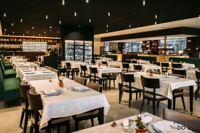 A large room with white tablecloths, green benches and a lit wall of wine bottles