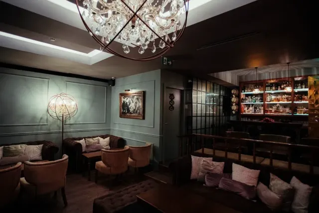 A dim bar lounge with a globe chandelier, velvet armchairs and lit bottle shelves