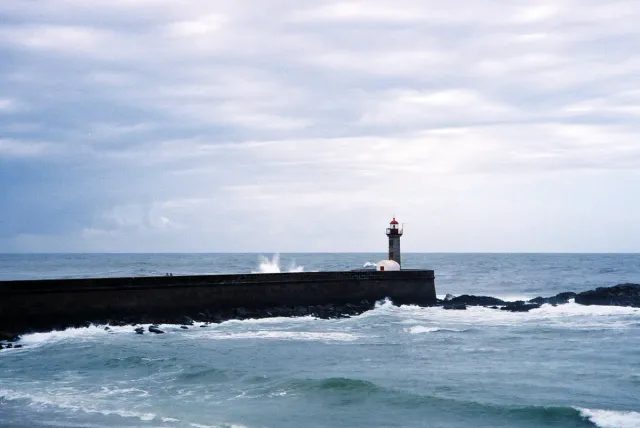 A small stone lighthouse at the end of a long pier with breaking waves