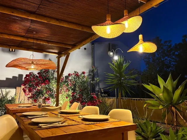 A bamboo pergola in the evening with straw lamps, a set table, flowering shrubs and an agave