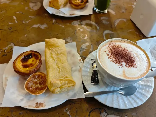 A marble table with a custard tart, a rolled pastry, a small cake and a cappuccino dusted with cocoa
