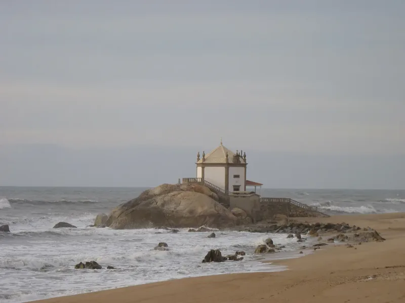Small white chapel perched on a rock in the sea beside sand and breaking waves