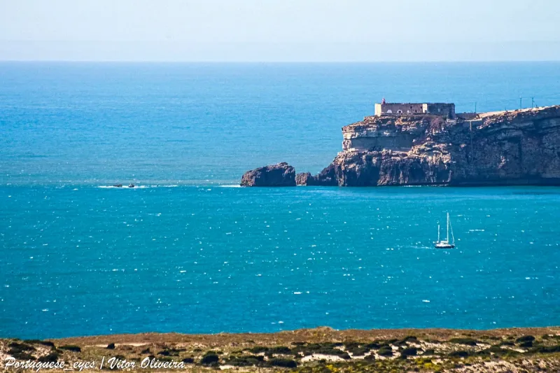 A fort and white village on top of a high cliff above turquoise sea with a sailing boat