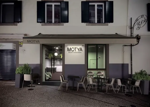A facade at night with a flat awning, a lit sign, chairs on the paving and a lit interior