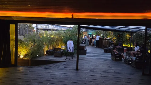 A wooden rooftop deck at dusk, with lit greenery and people sitting on benches