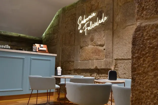 A stone wall with a handwritten neon sign, a light blue counter and pale armchairs around tables