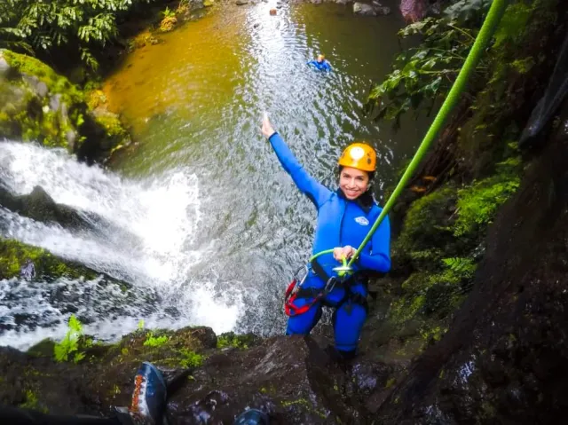 A woman in a blue suit and orange helmet holding a rope beside a canyon waterfall