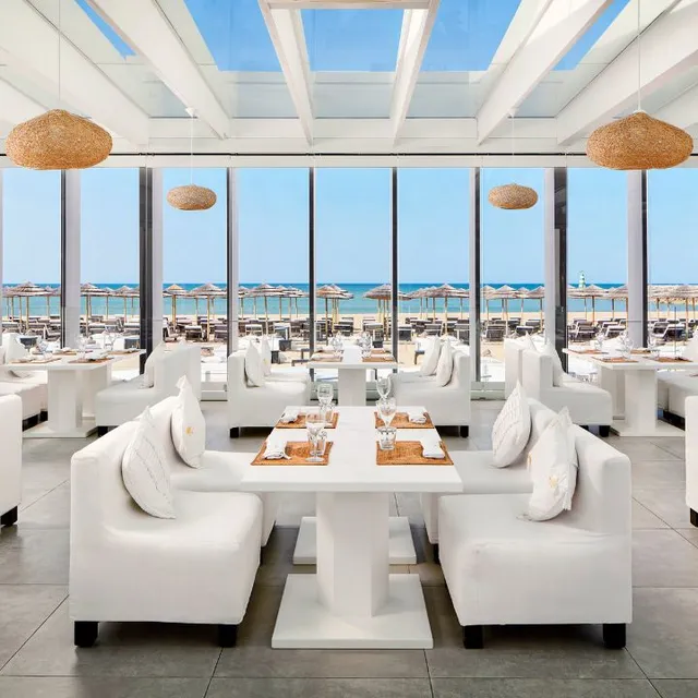 White sofas and set tables behind glass windows, facing a beach with straw umbrellas