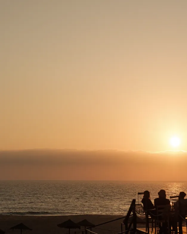 Silhouettes of people sitting on a terrace facing the sun setting over the ocean