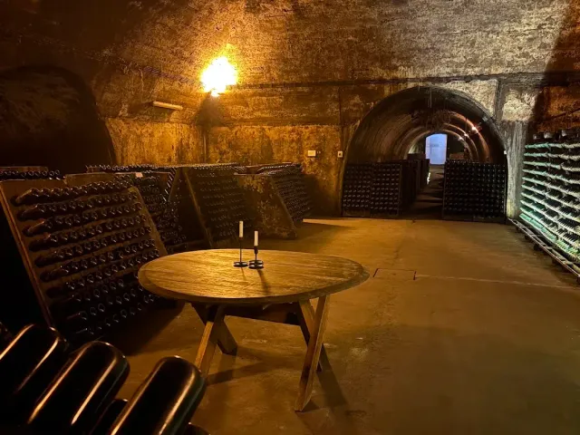 A vaulted underground cellar with stacks of bottles, a round wooden table and candles