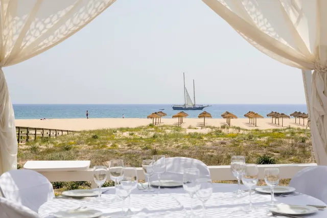 A set table under white curtains, facing a beach with straw umbrellas and a sailing ship