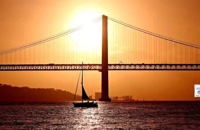 A silhouetted sailboat on a golden river at sunset, under a huge suspension bridge