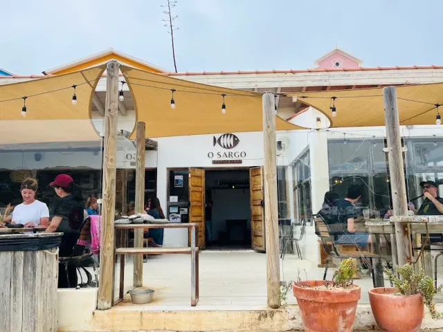 A yard shaded by fabric sails and string bulbs, diners around tables facing a white entrance