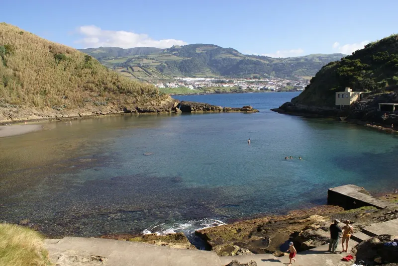 A round shallow lagoon inside a volcanic crater, with a narrow opening to the sea and green hills beyond