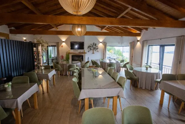 A hall with wooden beams and paper lamps, green chairs and set tables facing a window onto the water