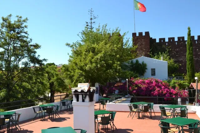 A terrace with green tables, a red fortress wall with a flag, and flowering bougainvillea