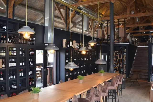 A long wooden table under hanging lamps, beside a shelf loaded with wine bottles