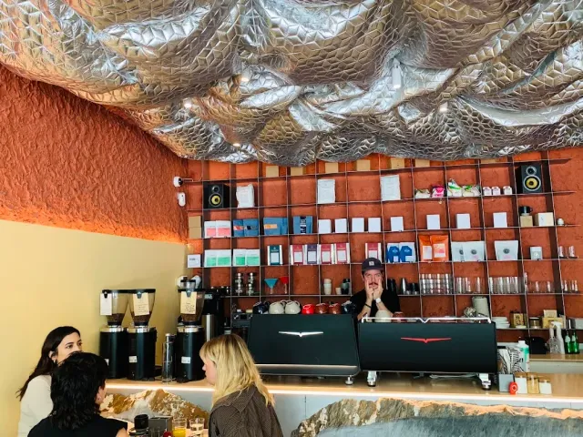 A barista behind an espresso machine, a shelf wall with coffee bags and a metal ceiling