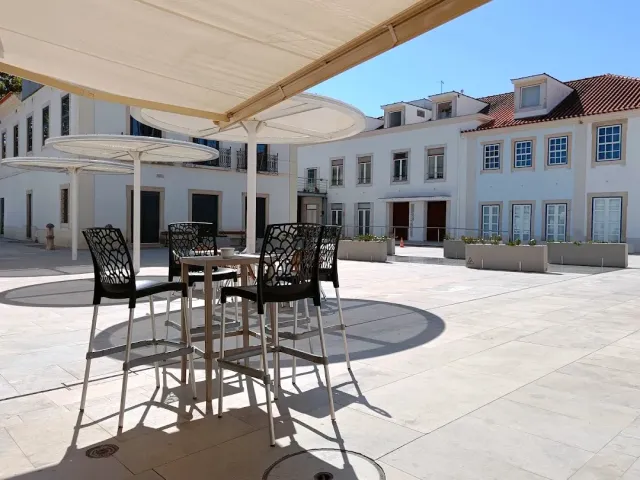 A paved square with a high table and black stools under an awning, facing white houses