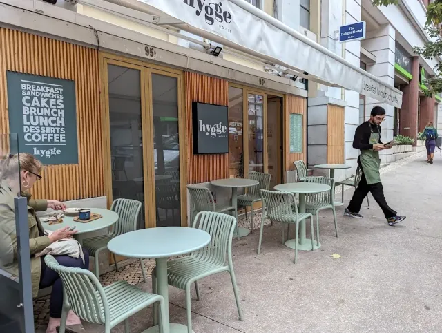 Pale green tables and chairs on a pavement in front of a wooden facade and a menu board