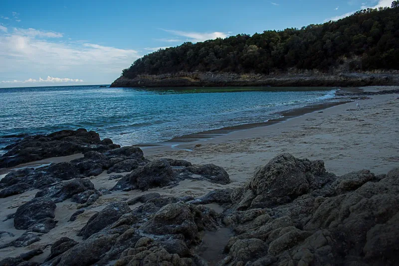 Shallow turquoise water and a strip of pale sand below a wooded cliff