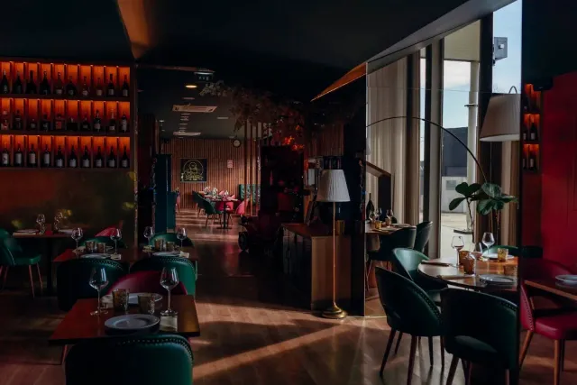 A dim room with green and red velvet chairs and lit wine shelves